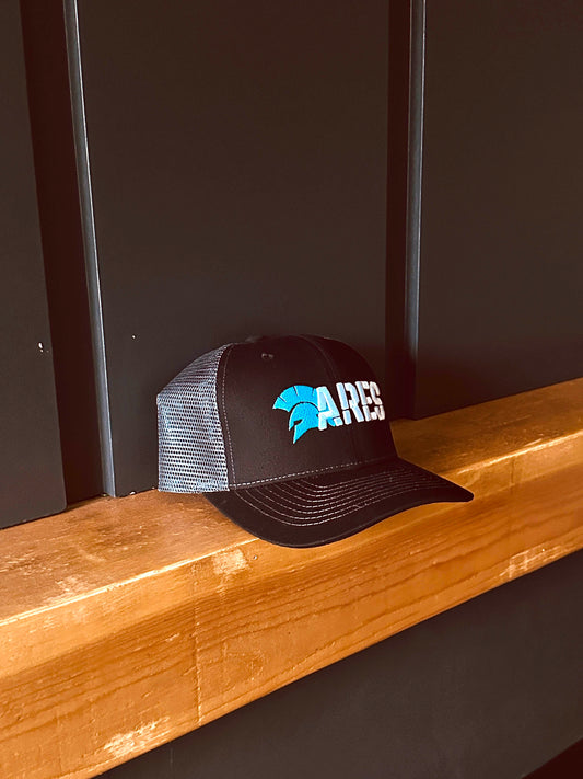 Black trucker hat with grey mesh and ARES logo on a wooden surface against a dark grey background